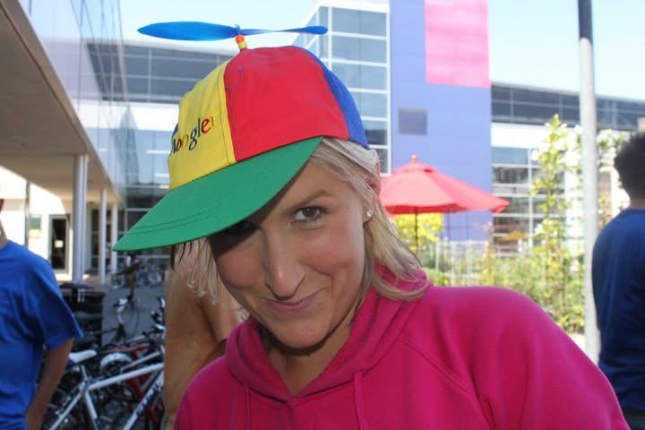 Woman wearing a colorful Google propeller hat and pink hoodie, smiling outside a modern office building with bikes and umbrellas.